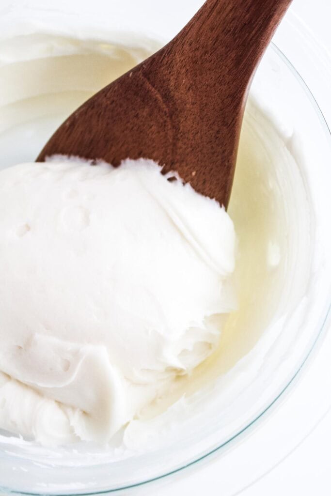 A large wooden spoon scooping cream cheese buttercream out of a bowl.